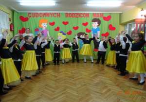 cDziewczynki w czarnych body i żółtych spódnicach oraz chłopcy ubrani na galowo śpiewają dla rodziców piosenkę. Dzieci mają uniesione w górę ręce.